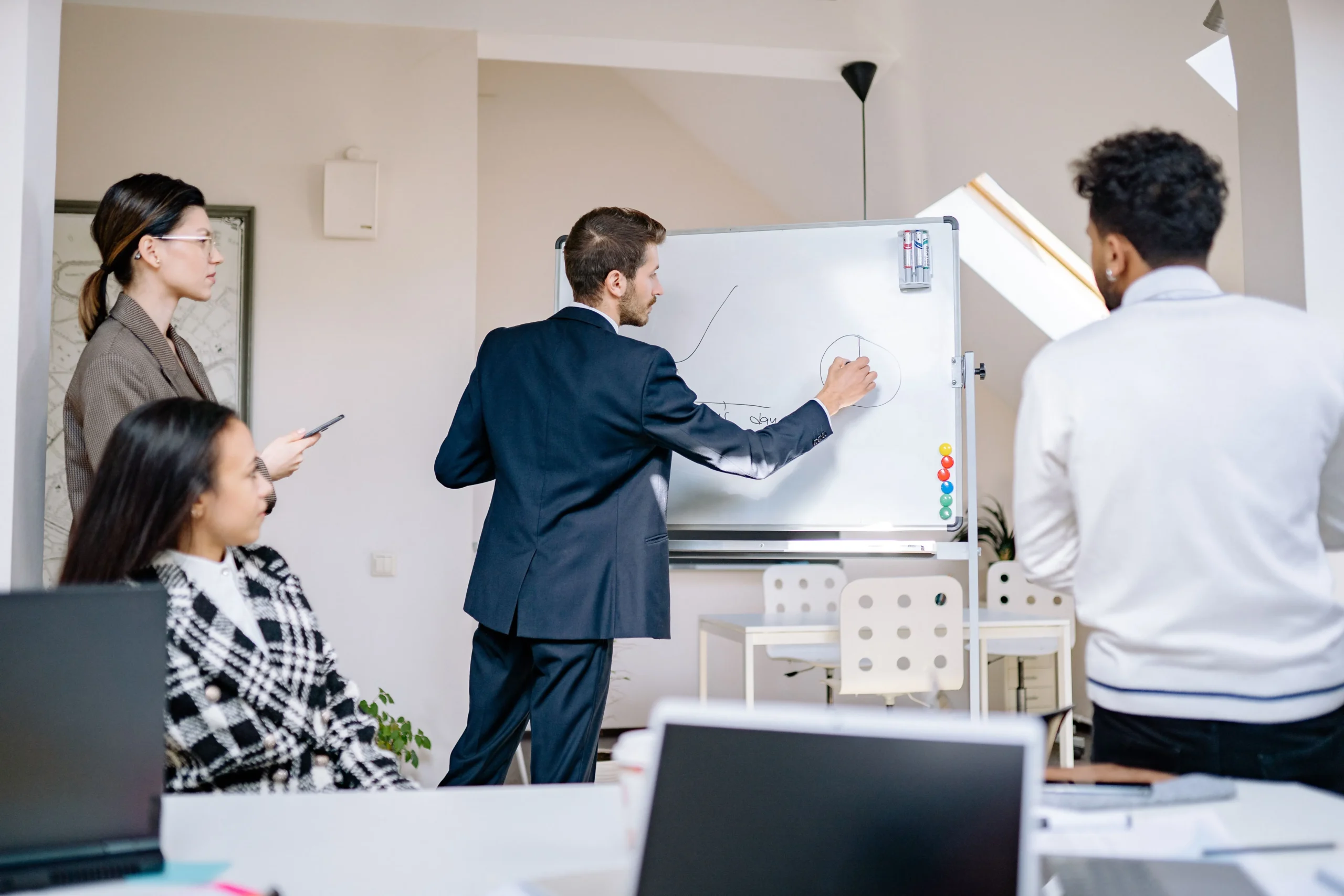Un homme en costume dessine un graphique de croissance sur un tableau blanc devant ses collaborateurs.
